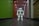A humanoid robot with white and gray accents stands in the middle of a hallway. The environment features a dark floor and a brightly lit ceiling with recessed lights. The walls are painted green on one side and light on the other, with various wooden panels and doors visible.