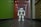 A humanoid robot with white and gray accents stands in the middle of a hallway. The environment features a dark floor and a brightly lit ceiling with recessed lights. The walls are painted green on one side and light on the other, with various wooden panels and doors visible.
