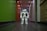 A humanoid robot with white and gray accents stands in the middle of a hallway. The environment features a dark floor and a brightly lit ceiling with recessed lights. The walls are painted green on one side and light on the other, with various wooden panels and doors visible.