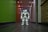 A humanoid robot with white and gray accents stands in the middle of a hallway. The environment features a dark floor and a brightly lit ceiling with recessed lights. The walls are painted green on one side and light on the other, with various wooden panels and doors visible.