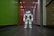 A humanoid robot with white and gray accents stands in the middle of a hallway. The environment features a dark floor and a brightly lit ceiling with recessed lights. The walls are painted green on one side and light on the other, with various wooden panels and doors visible.