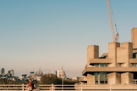 A modern concrete building stands prominently, with a construction crane extending skyward. In the background, a cityscape is visible, featuring an iconic dome and various skyscrapers. A person carrying a bag walks along the foreground, adding a human element to the urban environment.