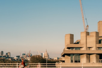 A modern concrete building stands prominently, with a construction crane extending skyward. In the background, a cityscape is visible, featuring an iconic dome and various skyscrapers. A person carrying a bag walks along the foreground, adding a human element to the urban environment.