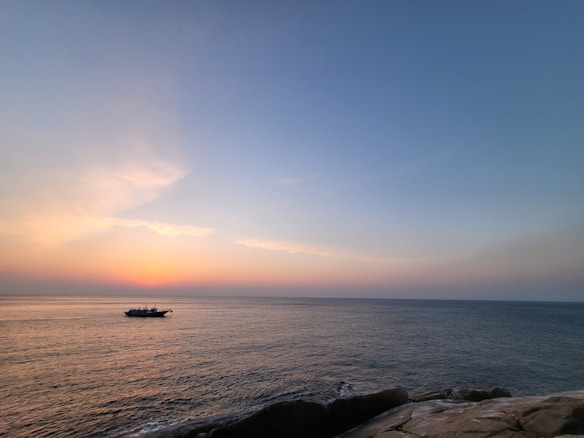 A serene ocean view at sunrise or sunset with a small boat on the horizon and gentle waves lapping against rocks in the foreground. The sky transitions from deep blue to warm hues of orange and pink.