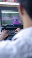 A person is using a laptop to read about ChatGPT, with a focus on the hands typing on the keyboard. The screen displays a webpage introducing ChatGPT, with text and graphical elements. The setting appears to be a casual or office environment, with soft lighting.