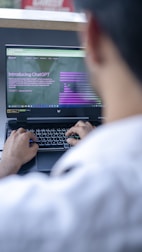 A person is using a laptop to read about ChatGPT, with a focus on the hands typing on the keyboard. The screen displays a webpage introducing ChatGPT, with text and graphical elements. The setting appears to be a casual or office environment, with soft lighting.