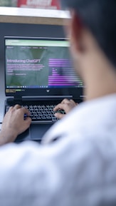 A person is using a laptop to read about ChatGPT, with a focus on the hands typing on the keyboard. The screen displays a webpage introducing ChatGPT, with text and graphical elements. The setting appears to be a casual or office environment, with soft lighting.