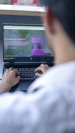 A person is using a laptop to read about ChatGPT, with a focus on the hands typing on the keyboard. The screen displays a webpage introducing ChatGPT, with text and graphical elements. The setting appears to be a casual or office environment, with soft lighting.