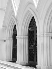 Gothic-style arched doorways with intricate design elements and dark metal doors are set against a backdrop of white stone walls. Footwear is placed on the entrance step, indicating a place of reverence or cultural significance.