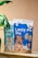 Two packages of pet milk substitute products are displayed, one for cats featuring a kitten, and one for dogs featuring a puppy. The packages are vibrant, with the cat product being blue and the dog product being blue and orange. A green plant leaf is partially visible on the left side of the image, with a soft beige background.