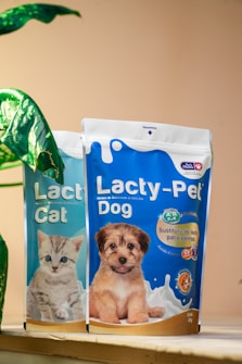 Two packages of pet milk substitute products are displayed, one for cats featuring a kitten, and one for dogs featuring a puppy. The packages are vibrant, with the cat product being blue and the dog product being blue and orange. A green plant leaf is partially visible on the left side of the image, with a soft beige background.