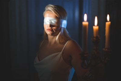 A woman wearing a blindfold stands in a dimly lit room. She is dressed in a white garment and holds a candelabrum with lit candles, casting a warm glow that contrasts with the deep shadows around her.