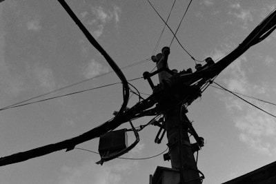 An overhead view of an electrical pole with multiple cables spread out in various directions. The pole is silhouetted against a cloudy sky, capturing the complexity of urban infrastructure.