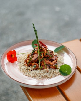A neatly arranged dish features a mound of seasoned meat garnished with herbs on a bed of rice. The plate is accented with vibrant red and green edible flowers, adding a touch of color. The setting includes a wooden surface, enhancing the rustic aesthetic.