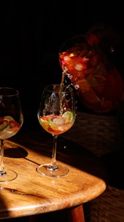 A pitcher is pouring a colorful fruit-infused beverage into a wine glass on a wooden table, with visible slices of strawberries and lime. The scene is warmly lit, casting sharp shadows and highlighting the vibrant colors of the drink.
