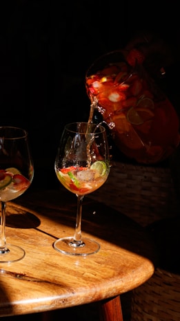 A pitcher is pouring a colorful fruit-infused beverage into a wine glass on a wooden table, with visible slices of strawberries and lime. The scene is warmly lit, casting sharp shadows and highlighting the vibrant colors of the drink.