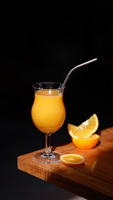 A glass filled with orange juice and a metal straw sits on a wooden table. Beside it, there are slices of fresh orange arranged attractively.