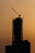 The silhouette of a high-rise building under construction is visible against a dusky, orange sky. A large crane is mounted on top of the structure, suggesting active development. The building's framework is clearly defined, emphasizing its skeletal design.