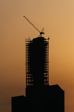 The silhouette of a high-rise building under construction is visible against a dusky, orange sky. A large crane is mounted on top of the structure, suggesting active development. The building's framework is clearly defined, emphasizing its skeletal design.