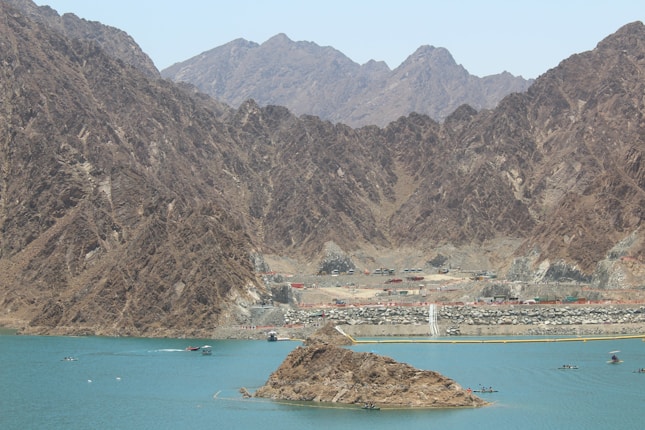 A vast and rugged mountain landscape with a clear blue lake in the foreground. The shoreline is rocky, and there are several small buildings along the base of the mountains. A few boats are visible on the water, adding a sense of activity to the otherwise serene setting.