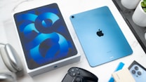 A blue iPad with an Apple logo is placed next to its box on a marble surface. Surrounding the iPad are various gadgets including headphones, a game controller, and a smartphone. There is also a plant in a white pot, contributing to a modern and tech-savvy setting.