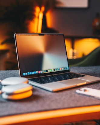 A sleek laptop on a desk with its screen displaying a colorful gradient wallpaper. Next to the laptop, there are wireless headphones and a smartphone. The background features warm ambient lighting and blurred decor elements.
