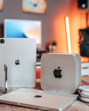 A close-up shot of various Apple products including a Mac Mini, iPad Pro, iPhone, and MacBook on a wooden table. The background is softly blurred and features warm lighting.