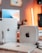 A close-up shot of various Apple products including a Mac Mini, iPad Pro, iPhone, and MacBook on a wooden table. The background is softly blurred and features warm lighting.