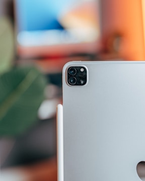 A close-up view of the back of a silver tablet featuring dual camera lenses and a flash in the top corner, along with a stylus attached to its side. A blurred background showcases a colorful screen and green foliage, creating a modern and tech-focused ambiance.