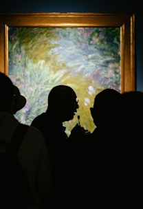 A group of people is gathered in front of a framed artwork, with the focus on a person holding a microphone in silhouette against the backdrop of the painting. The painting appears to be an impressionistic piece, mainly with tones of green, blue, and hints of purple.