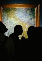A group of people is gathered in front of a framed artwork, with the focus on a person holding a microphone in silhouette against the backdrop of the painting. The painting appears to be an impressionistic piece, mainly with tones of green, blue, and hints of purple.