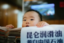 A baby peeks over the edge of a surface with wide eyes. The background is blurred, featuring warm tones and a screen displaying a colorful image. In the foreground, there is a piece of fabric or paper with Chinese characters printed on it.