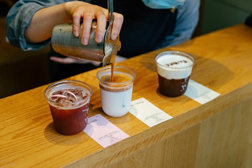 A person is pouring a beverage from a stainless steel pitcher into a plastic cup. There are three plastic cups on a wooden counter, each filled with a different drink. One cup contains a dark iced beverage, another has a white creamy drink topped with a layer of brown liquid, and the third is filled with a frothy dark drink.