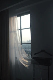 A serene shot of a charcoal black tailored coat hanging by a sunlit window with sheer white curtains.