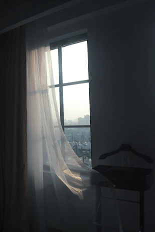 A serene shot of a charcoal black tailored coat hanging by a sunlit window with sheer white curtains.