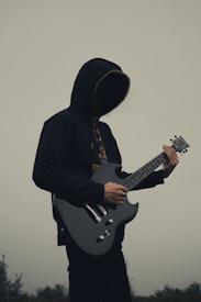 A person with a hooded sweatshirt playing an electric guitar. The background is a muted, overcast sky, creating a mysterious or somber atmosphere. The person's face is obscured by the hood, adding an element of anonymity or intrigue.