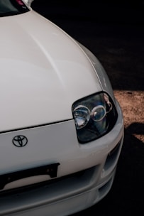 A close-up view of the front end of a white car featuring a prominent headlight and the Toyota logo. The car's sleek design and glossy finish are highlighted, with shadows accentuating its curves.