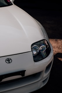 A close-up view of the front end of a white car featuring a prominent headlight and the Toyota logo. The car's sleek design and glossy finish are highlighted, with shadows accentuating its curves.