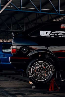 A black car with visible racing decals and advertisements is elevated on a red jack stand in a garage or workshop. The well-polished tire shows 'Hoosier' branding and elaborate wheel design. A blue vehicle is parked in the background under the metal framework of the garage roof.