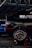 A black car with visible racing decals and advertisements is elevated on a red jack stand in a garage or workshop. The well-polished tire shows 'Hoosier' branding and elaborate wheel design. A blue vehicle is parked in the background under the metal framework of the garage roof.