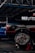 A black car with visible racing decals and advertisements is elevated on a red jack stand in a garage or workshop. The well-polished tire shows 'Hoosier' branding and elaborate wheel design. A blue vehicle is parked in the background under the metal framework of the garage roof.