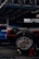 A black car with visible racing decals and advertisements is elevated on a red jack stand in a garage or workshop. The well-polished tire shows 'Hoosier' branding and elaborate wheel design. A blue vehicle is parked in the background under the metal framework of the garage roof.