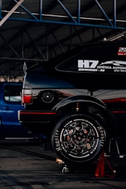 A black car with visible racing decals and advertisements is elevated on a red jack stand in a garage or workshop. The well-polished tire shows 'Hoosier' branding and elaborate wheel design. A blue vehicle is parked in the background under the metal framework of the garage roof.
