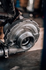 A close-up of a metallic turbocharger, showcasing intricate engineering with bolts and flanges. The machinery appears sturdy and robust, highlighted by a background of blurred mechanical components.