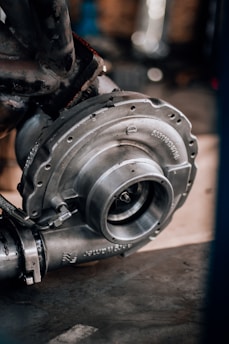 A close-up of a metallic turbocharger, showcasing intricate engineering with bolts and flanges. The machinery appears sturdy and robust, highlighted by a background of blurred mechanical components.