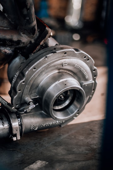 A close-up of a metallic turbocharger, showcasing intricate engineering with bolts and flanges. The machinery appears sturdy and robust, highlighted by a background of blurred mechanical components.