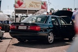 A black sedan is parked with both the back door and trunk open. In the background, there is a booth or stall with a sign featuring strawberries and the name 'Moranginha Sta Cruz'. The scene appears to be in an outdoor area with other people and objects partially visible.