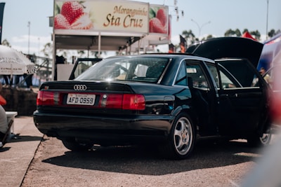 A black sedan is parked with both the back door and trunk open. In the background, there is a booth or stall with a sign featuring strawberries and the name 'Moranginha Sta Cruz'. The scene appears to be in an outdoor area with other people and objects partially visible.