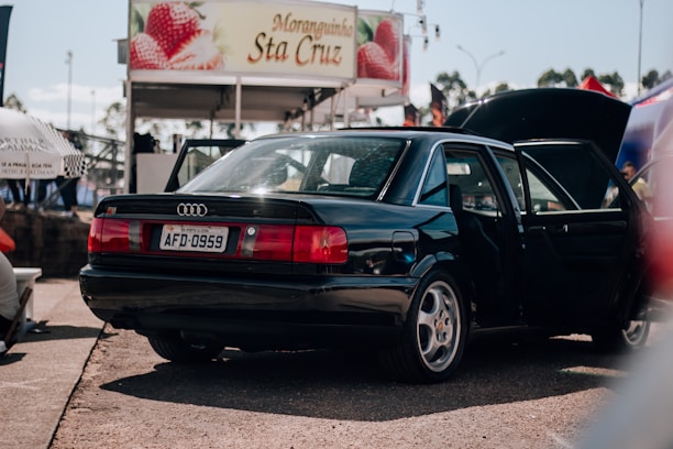 A black sedan is parked with both the back door and trunk open. In the background, there is a booth or stall with a sign featuring strawberries and the name 'Moranginha Sta Cruz'. The scene appears to be in an outdoor area with other people and objects partially visible.