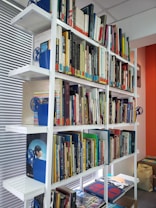 A well-organized bookshelf filled with a diverse collection of books arranged in various colors and sizes. The shelves are white and made of metal, contributing to a clean and modern look. Various blue bookends with a globe design are used to support the books, and the wall beside the shelf is painted in a warm, orange tone.